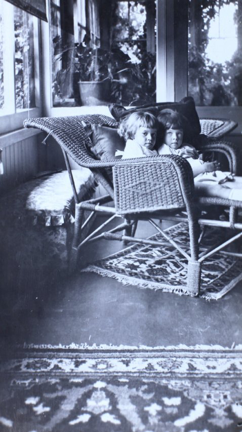 Two small girls sitting on a cane lounge in a sunroom