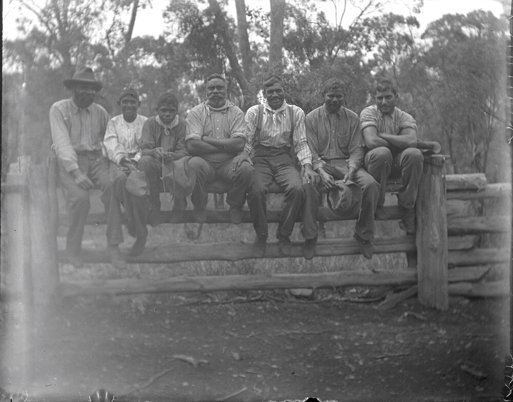 Seven Aboriginal stockmen sitting on post and rail fence