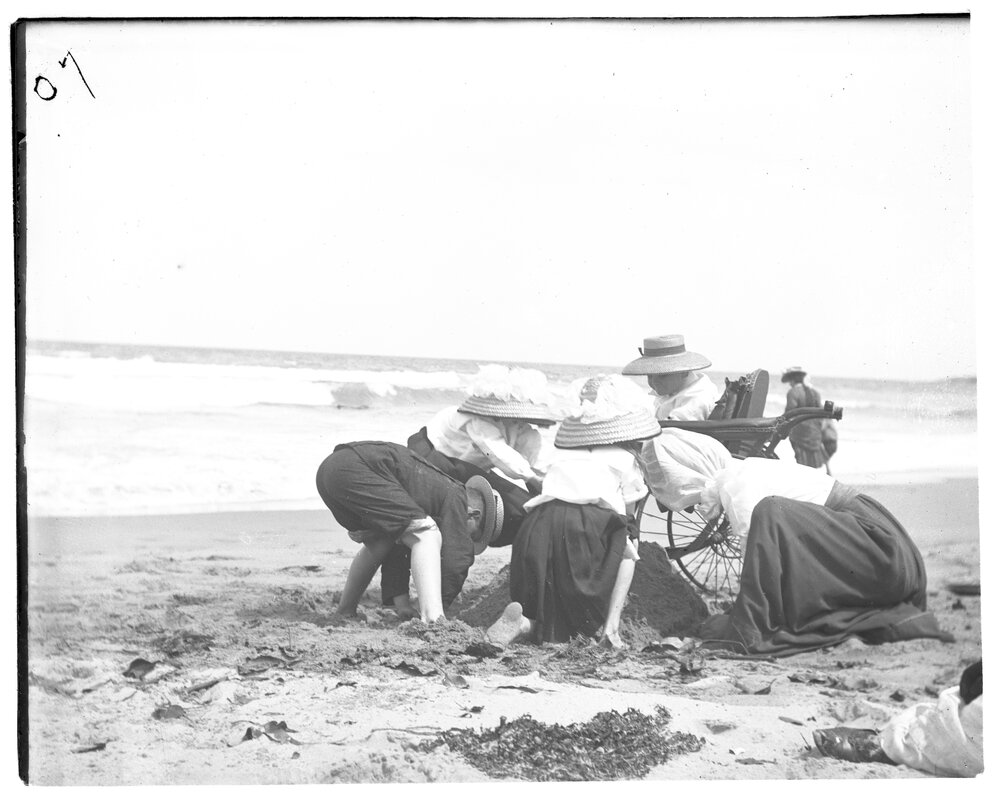 Children playing in the sand beside a woman in a wheelchair at the beach, 1907
