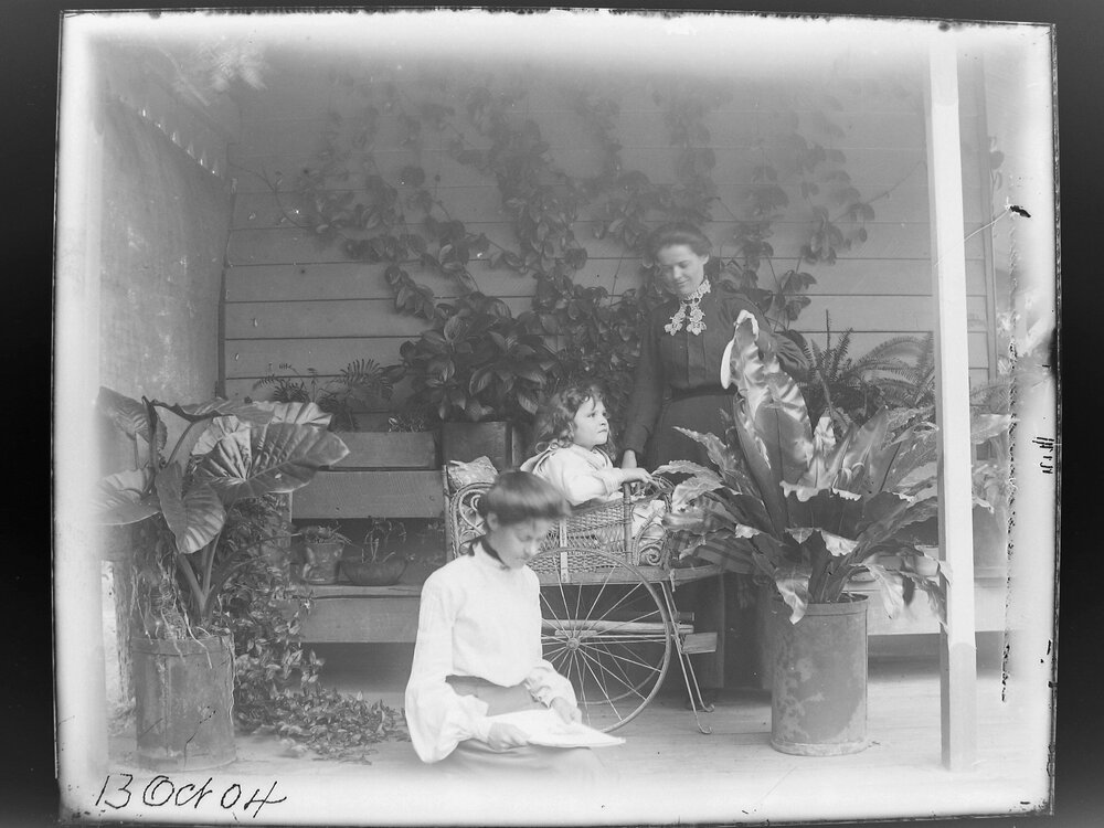 Two women and a child on a verandah with potted plants, 1904