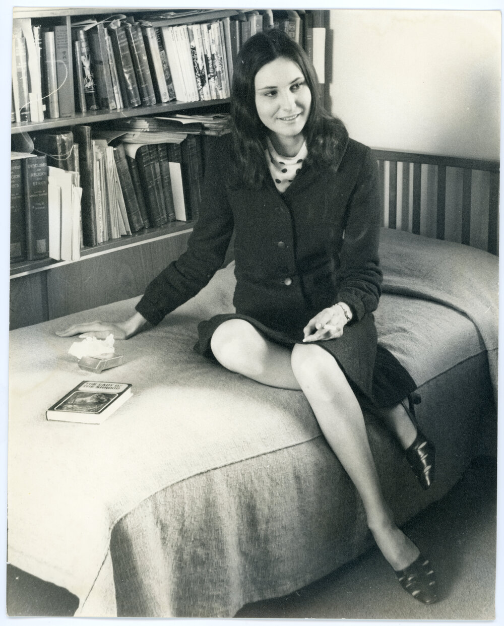 Female student seated on bed in front of bookshelf with The Lady of the Shroud, cigarette packet and book