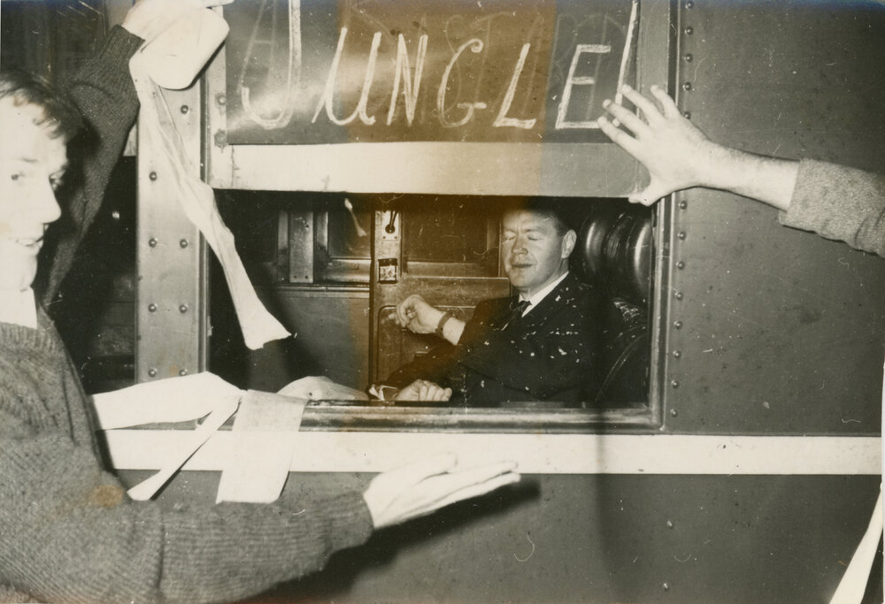 Man viewed through train window with Wright College students outside draping the train in toilet paper, 1964