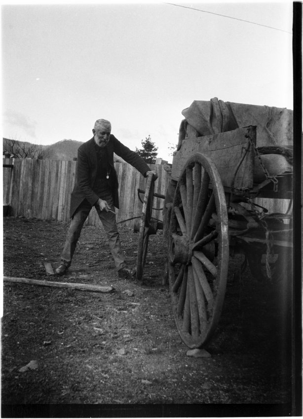 Unidentified older man standing by a wooden cart