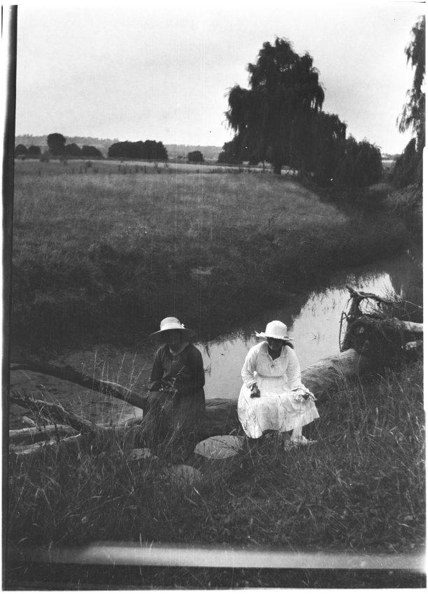Two unidentified women sitting of a log by a creek
