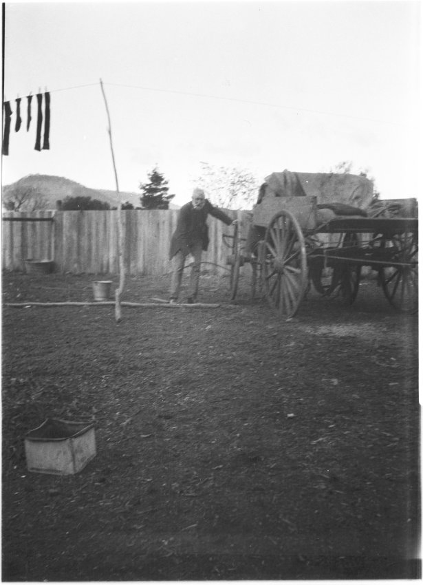 Unidentified older man standing by a wooden cart