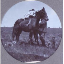 Unidentified woman on horseback with a man and pet dogs