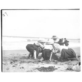 Children playing in the sand beside a woman in a wheelchair at the beach, 1907