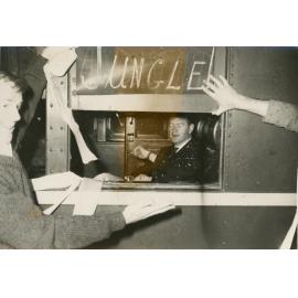 Man viewed through train window with Wright College students outside draping the train in toilet paper, 1964