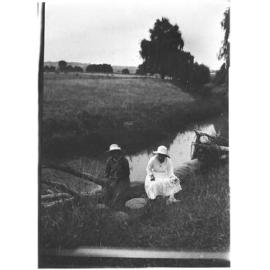 Two unidentified women sitting of a log by a creek
